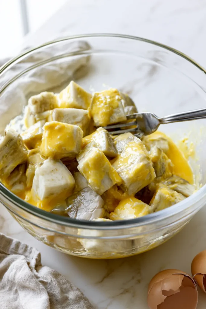 Peeled eggplant cubes coated in beaten egg in a glass mixing bowl with a fork, eggshells beside the bowl