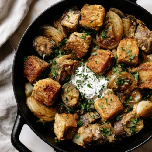 Golden egg-coated eggplant cubes with soft onion wedges and chopped parsley in a cast iron skillet, a spoonful of sour cream in the center