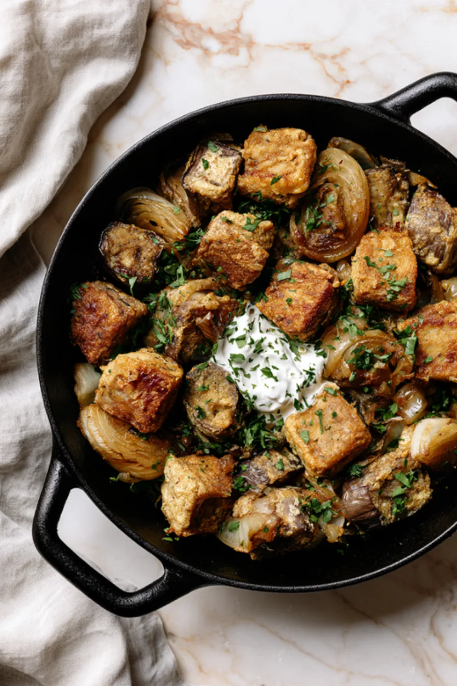 Golden egg-coated eggplant cubes with soft onion wedges and chopped parsley in a cast iron skillet, a spoonful of sour cream in the center