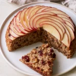 Flourless oatmeal apple cake topped with a fan of glazed apple slices, one slice cut to show the oat crumb with walnuts and cranberries