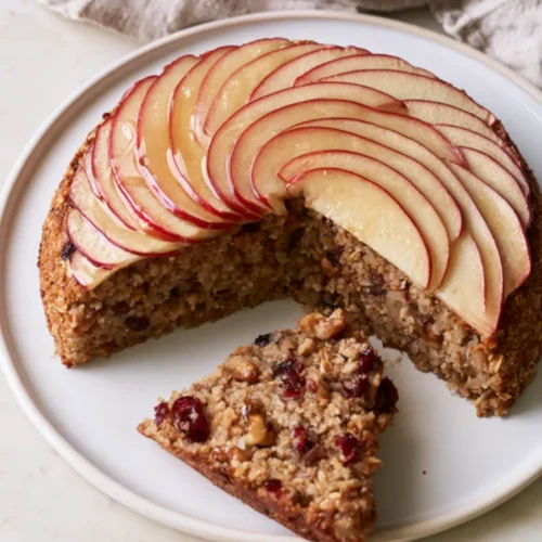 Flourless oatmeal apple cake topped with a fan of glazed apple slices, one slice cut to show the oat crumb with walnuts and cranberries