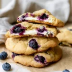 Stack of gluten-free blueberry cheesecake cookies on parchment, the top two broken open to show a creamy cream cheese center and burst blueberries