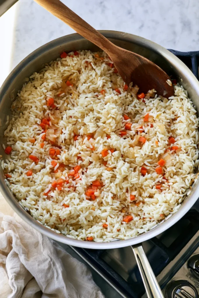 Toasting the Rice with Onion and Carrots Long-grain rice stirred into softened onion and diced carrots in a stainless steel skillet, with a wooden spoon on the rim